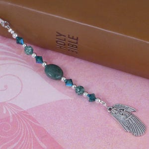 May include: A green and silver beaded bookmark with an angel charm. The bookmark is on top of a brown leather-bound book with the words "HOLY BIBLE" embossed on the cover.