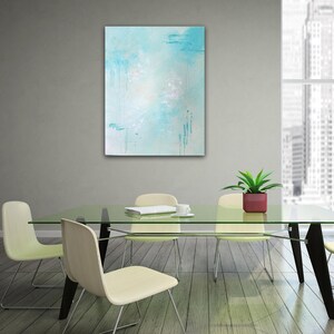 May include: A light blue abstract painting with white and light blue accents hangs on a wall above a glass table with four chairs. The table is set with a magazine and a cup.