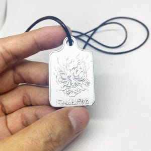 Może przedstawiać: Srebrny prostokątny wisiorek z wygrawerowanym wizerunkiem maski samuraja z płomieniami. Słowo "SAMURAI" jest wyryte na dole. Wisiorek zawieszony jest na czarnym sznurku.