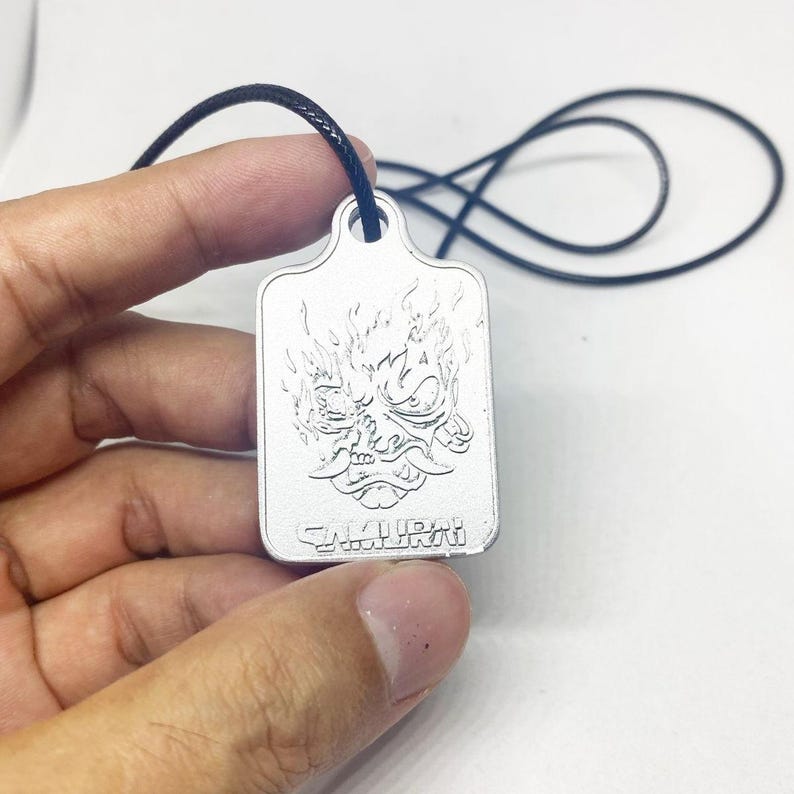 Może przedstawiać: Srebrny prostokątny wisiorek z wygrawerowanym wizerunkiem maski samuraja z płomieniami. Słowo "SAMURAI" jest wyryte na dole. Wisiorek zawieszony jest na czarnym sznurku.