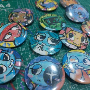 May include: A collection of colorful, cartoon-style button pins. Each pin features a unique character design with vibrant colors, including blues, yellows, and pinks. The pins have a glossy finish and are arranged on a green surface.