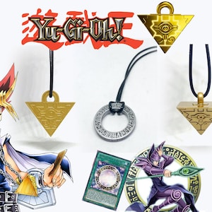 May include: Image showing Yu-Gi-Oh! themed pendants and a card. The pendants are gold and silver, with different shapes and designs. The Yu-Gi-Oh! logo is in red. The card has a purple and white design. The background is white.