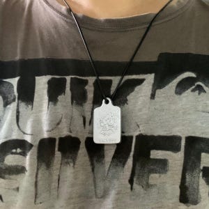 Może przedstawiać: Srebrny prostokątny wisiorek z ozdobnym wzorem wisi na czarnym sznurkowym naszyjniku. Wisiorek zawieszony jest na szarej koszulce z dużym, wyblakłym czarnym nadrukiem.