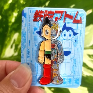 May include: Astro Boy pin badge with a split design: one side shows the character's exterior, the other reveals internal mechanics. The badge is on a blue background with Japanese text and a cartoon illustration of Astro Boy.