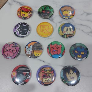 Pode incluir: Uma coleção de doze botões redondos coloridos com desenhos de personagens de desenhos animados. Os botões apresentam várias ilustrações em cores brilhantes, incluindo rostos, logotipos e figuras. Os botões estão dispostos em uma superfície clara.