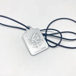 Może przedstawiać: Srebrny prostokątny wisiorek z szczeg&oacute;łowym grawerowanym wzorem twarzy demona z płomieniami i napisem "SAMURAI" na dole. Wisiorek zawieszony jest na czarnym sznurku. Tło jest białe.