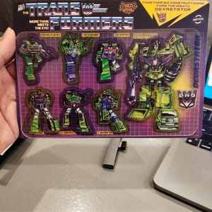 May include: A vintage Transformers sticker sheet featuring the Constructicon characters. The sheet includes individual stickers of Bonecrusher, Scavenger, Scrapper, Long Haul, Mixmaster, and Devastator. The packaging has the text "More Than Meets The Eye!"
