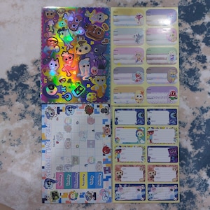 Könnte beinhalten: Eine Sammlung von Schulmaterialien mit Zeichentrickfiguren. Enthält ein holografisches Aufkleberblatt, Namensschilder und einen Stundenplan. Der Stundenplan hat Wochentage und ein Raster zur Planung. Die Namensschilder haben Platz für Name und Fach.
