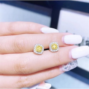 May include: A pair of square-shaped stud earrings. Each earring features a central yellow stone surrounded by a halo of smaller, clear stones. The earrings are set in a silver-colored metal and are displayed on a light-toned background.
