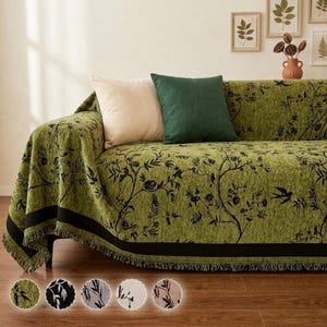 May include: A green sofa cover featuring a black botanical print of branches, leaves, and birds. The cover has a solid black horizontal stripe near the bottom edge and a fringed hem. It is styled with one cream and one dark green square pillow.