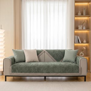 May include: A three-seat sofa featuring a light beige frame and dark green textured fabric covers on the seat and backrest. The sofa includes two light green square throw pillows and one beige square throw pillow, all with a soft fabric finish.