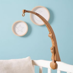 May include: Wooden baby mobile arm in a natural finish, attached to a white crib. The curved arm has a hook for hanging mobiles. Two circular wall decorations with a star and moon are visible.