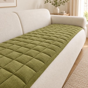 Peut inclure: Une housse de canapé vert olive rectangulaire présentant un motif carré matelassé avec une texture à chevrons subtile. La housse est posée à plat sur l'assise d'un canapé crème, offrant une couche protectrice au design en grille uniforme.