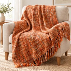 Könnte beinhalten: Eine gewebte Wohndecke mit einem mehrfarbigen Muster aus leuchtend orangefarbenen, rostbraunen und cremefarbenen Fäden. Das rechteckige Textil hat ein strukturiertes, grobes Gewebe und ist am unteren Rand mit langen, geknoteten Fransen versehen.