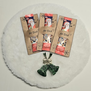 Może przedstawiać: Trzy czekoladowe smakołyki w czerwono-niebieskich opakowaniach, każdy z motywem Świętego Mikołaja, prezentowane są na brązowych kartach z napisem "für Dich". Karty zdobione są płatkami śniegu i ilustracjami bałwanków. Dwa zielone dzwoneczki zdobią dół.