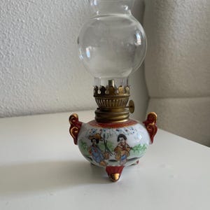 Könnte beinhalten: Eine kleine, antike Öllampe mit einem durchsichtigen Glaskolben und einem dekorativen Keramiksockel. Der Sockel zeigt eine bemalte Szene mit Figuren in traditioneller Kleidung, akzentuiert mit roten und goldenen Details. Die Lampe hat einen Messingbrenner.