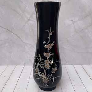 Pode incluir: Um vaso preto com a parte superior alargada e base redonda. O vaso é decorado com um desenho floral e de pássaros em madrepérola. O desenho inclui flores, folhas e pássaros pousados em galhos.