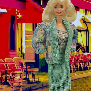 May include: A doll wearing a green knit skirt suit with a matching jacket. The jacket has a floral pattern on the sleeves. The doll has blonde hair and is standing in front of a cafe scene.