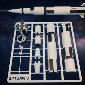 May include: A white and black Saturn V model rocket kit with various components on a white plastic frame. The assembled rocket is at the top, with "USA" printed on the side. The frame has the text "SATURN V".