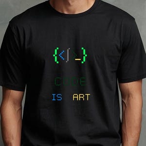 Könnte beinhalten: Schwarzes T-Shirt mit dem Text "CODE IS ART" in pixeliger grüner, blauer und gelber Schrift. Das Shirt zeigt ein pixeliges Codesymbol über dem Text. Das Model trägt eine weiße Hose.