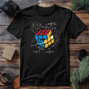 Pode incluir: Camiseta preta com um gráfico de um Cubo de Rubik e equações matemáticas, incluindo "Π= 3.14". O cubo é vermelho, azul e amarelo. O design inclui setas brancas e fórmulas manuscritas.