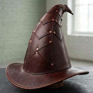 May include: A brown leather witch hat with a wide brim and a pointed crown. The hat features decorative stitching and copper-colored rivets. The hat is designed for costume or cosplay.