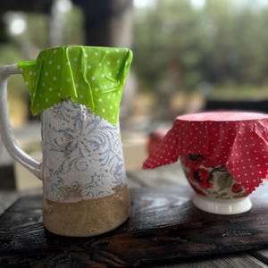 Puede incluir: Dos tazas de cerámica, una alta con asa y diseño floral, la otra una taza de té más pequeña. Ambas están cubiertas con envoltorios de alimentos reutilizables. La taza alta tiene una envoltura verde con lunares blancos. La taza de té tiene una envoltura roja con lunares blancos.