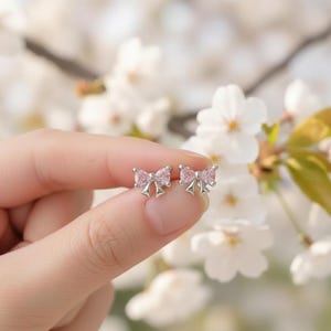 May include: A pair of silver bow-shaped stud earrings with pink gemstone centers. The earrings are held in a hand against a backdrop of white flowers, showcasing the delicate design and color.