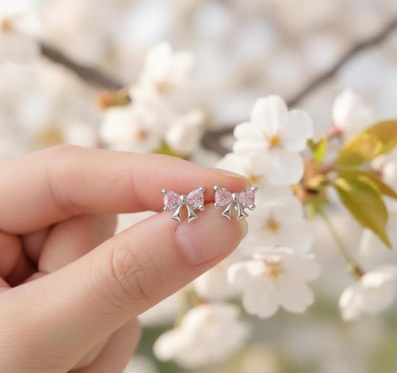 May include: A pair of silver bow-shaped stud earrings with pink gemstone centers. The earrings are held in a hand against a backdrop of white flowers, showcasing the delicate design and color.