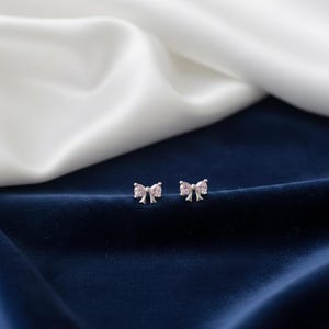 May include: A pair of silver bow-shaped stud earrings with pink gemstone accents. The earrings are displayed on a dark blue velvet surface with a white and off-white satin background.