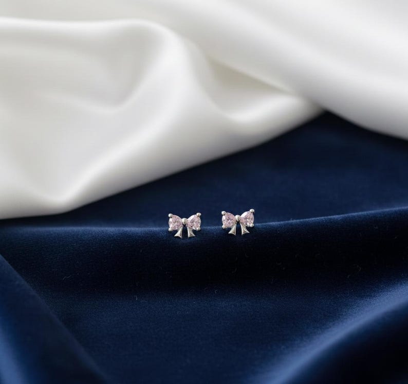 May include: A pair of silver bow-shaped stud earrings with pink gemstone accents. The earrings are displayed on a dark blue velvet surface with a white and off-white satin background.