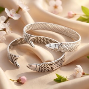 May include: Two silver-toned cuff bracelets shaped like fish. Each bracelet features a detailed scale pattern along the band, with a fish head at one end and a tail fin at the other. The metal has a polished finish with darkened recessed areas to highlight the texture of the scales and the facial features of the fish.