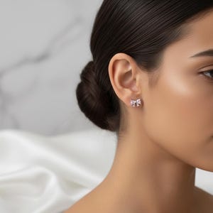 May include: A close-up of a person's ear with a small, bow-shaped stud earring. The earring is silver-toned with a pink gemstone center. The background is blurred, with a white fabric and a marble-like surface.