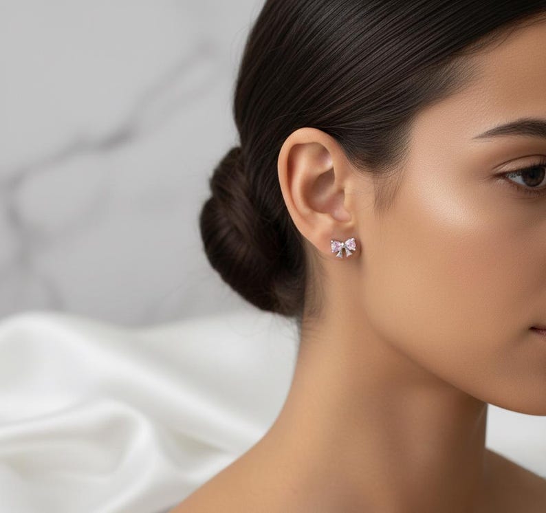 May include: A close-up of a person's ear with a small, bow-shaped stud earring. The earring is silver-toned with a pink gemstone center. The background is blurred, with a white fabric and a marble-like surface.