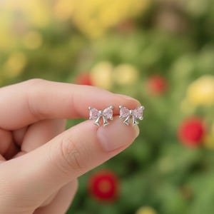 May include: A pair of silver bow-shaped stud earrings with pink gemstone accents. The earrings are held in a hand against a blurred green and yellow background.
