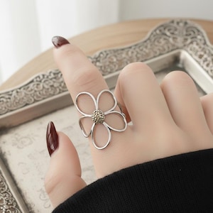 Może przedstawiać: Srebrny pierścionek w kształcie kwiatu z pięcioma płatkami. Środek kwiatu ma teksturowany, złoty wzór. Pierścionek jest noszony na palcu, a tło stanowi dekoracyjna, srebrna taca.