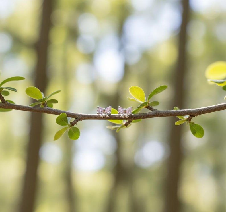May include: A pair of silver butterfly earrings with pink gemstones, resting on a slender tree branch with green leaves. The background is blurred, suggesting an outdoor setting.