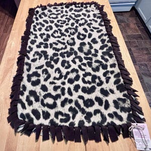 May include: A rectangular fleece blanket featuring a black and white leopard print pattern. The edges of the blanket are finished with a knotted fringe border made of solid dark brown fabric strips. A small white product tag is attached to one corner.