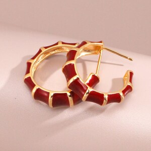 May include: Gold-coloured hoop earrings with a bamboo-style design. The hoops are detailed with a rich, glossy red enamel, creating a striking contrast. The earrings are presented on a soft, neutral background.