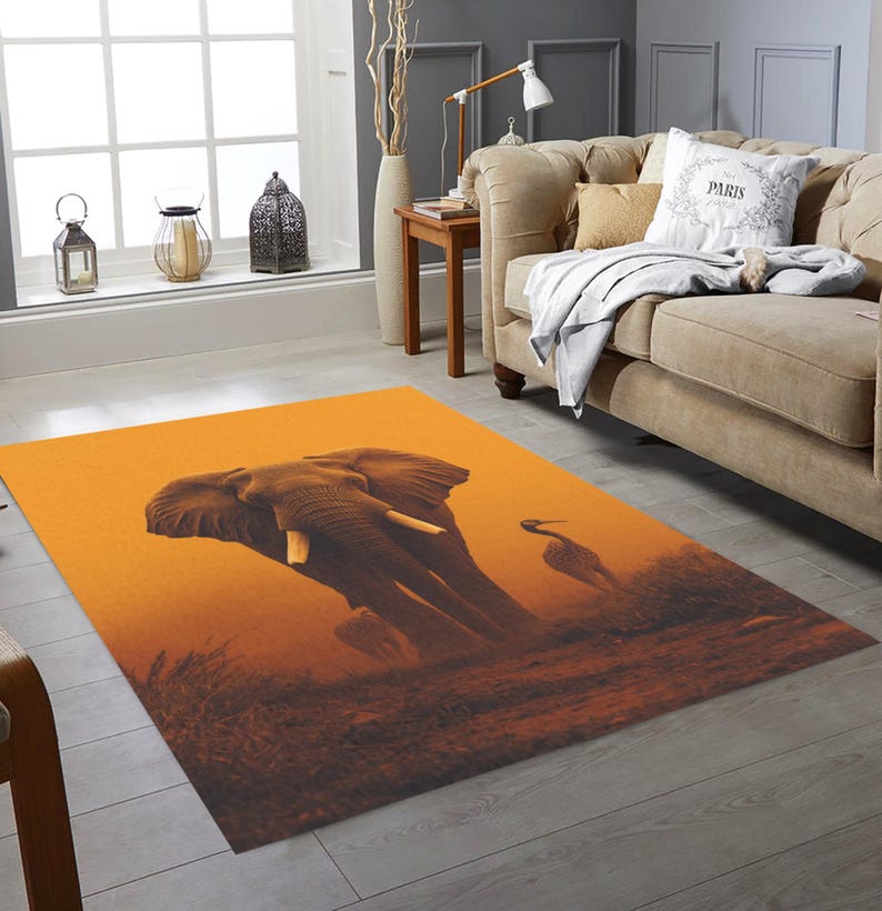 May include: An orange area rug featuring an elephant and a bird silhouette against a sunset backdrop. The rug has a rectangular shape and is placed in a living room setting, near a sofa and window.