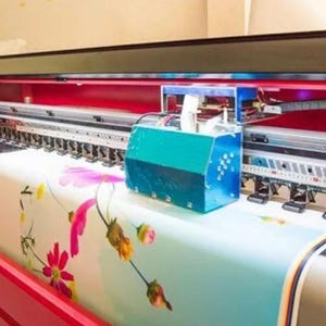 May include: A large format printer in red and black, printing a floral pattern on fabric. The machine has a teal box attached and is in an indoor setting. The fabric being printed has pink and yellow flowers.