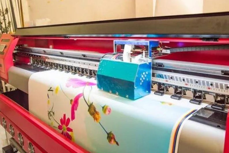 May include: A large format printer in red and black, printing a floral pattern on fabric. The machine has a teal box attached and is in an indoor setting. The fabric being printed has pink and yellow flowers.