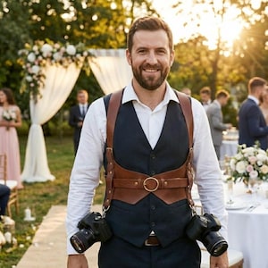 Pode incluir: Um fotógrafo usando um arnês de câmera de couro marrom com duas câmeras pretas. Ele está vestindo uma camisa branca, um colete azul marinho e está sorrindo. O fundo mostra uma cerimônia de casamento.