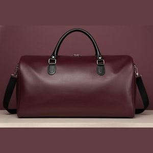May include: A burgundy leather duffel bag with black handles and a black shoulder strap. The bag has a rectangular shape and a zip closure. The handles are curved and attached with silver-coloured hardware.
