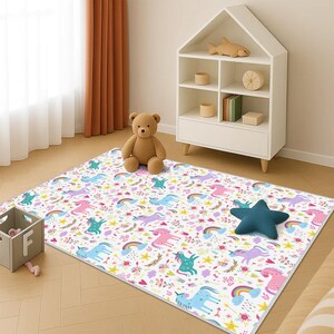 Può includere: Un tappeto rettangolare bianco con un motivo colorato di unicorni, arcobaleni e stelle. Il tappeto presenta unicorni rosa, blu e viola. Un orsacchiotto marrone e un cuscino blu a forma di stella sono sul tappeto.