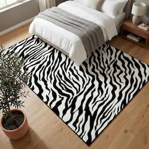 May include: A large rectangular rug with a zebra stripe pattern in black and white. The rug is placed on a hardwood floor in a bedroom setting, partially under a bed with white bedding.