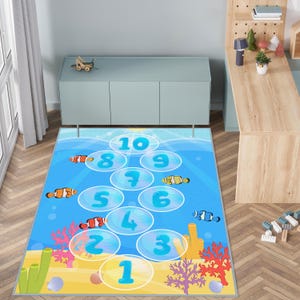 May include: A rectangular children's play rug featuring a hopscotch design with numbers 1-10 in blue bubbles. The rug has an underwater theme with cartoon fish, coral, and a sandy seabed in shades of blue, yellow, and orange.