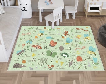 Op de afbeelding: Lichtgroen kinderkleed met alfabet en insectenillustraties. Het kleed heeft een wit stippenpatroon en toont mieren, bijen, vlinders en spinnen. Het kleed ligt in een kinderkamer, met een tafel en stoelen.