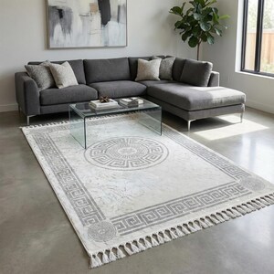 May include: A large, rectangular area rug with a Greek key pattern in shades of gray and white. The rug features a central medallion design and a fringed edge. The rug is placed in a living room setting with a gray sectional sofa and glass coffee table.