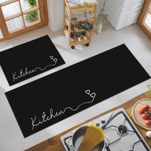 Puede incluir: Alfombras de cocina negras con texto blanco que dice "Kitchen" y diseños de corazones. Una alfombra es rectangular y la otra cuadrada. Las alfombras están sobre un suelo blanco, cerca de una estufa y un carrito de madera.